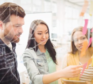 Three co-workers putting sticky notes on clear wall in office
