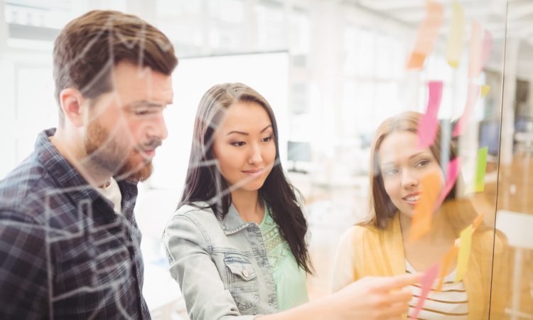 Three co-workers putting sticky notes on clear wall in office