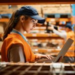 Female warehouse worker working on laptop at storage compartment.