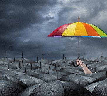 Sea of black umbrellas amid storm with hand holding rainbow umbrella above the rest