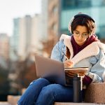 Young woman writing in notebook with laptop on lap sitting on stairs outside on university campus
