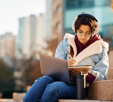 Young woman writing in notebook with laptop on lap sitting on stairs outside on university campus