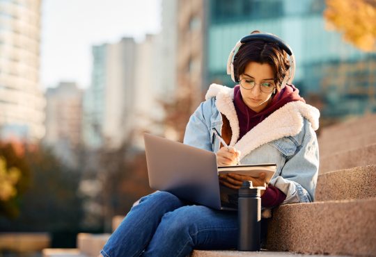 Young woman writing in notebook with laptop on lap sitting on stairs outside on university campus