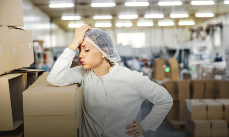 Woman wearing white lab coat and hairnet in factory leaning against stack of boxes with head in hand