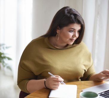 Woman working on computer and writing in notebook at home