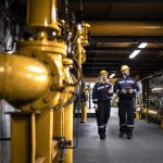 Industrial workers in protective work wear walking by gas pipeline inside refinery and checking oil production.