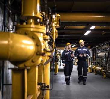 Industrial workers in protective work wear walking by gas pipeline inside refinery and checking oil production.