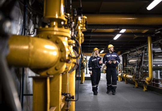 Industrial workers in protective work wear walking by gas pipeline inside refinery and checking oil production.