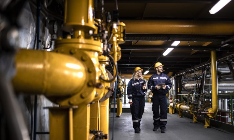 Industrial workers in protective work wear walking by gas pipeline inside refinery and checking oil production.