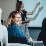 Rear view of a woman sitting in audience looking back at a colleague and smiling during a conference.