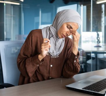 Stressed woman sitting at desk, leaning head down, gripping bridge of nose between thumb and forefinger