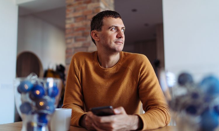 A man wearing yellow sweater sitting in the kitchen and looking through window while using smartphone