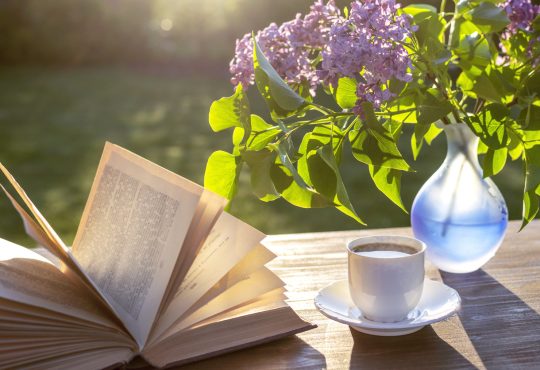 Open book sitting on table outdoors beside teacup and vase with purple lilacs