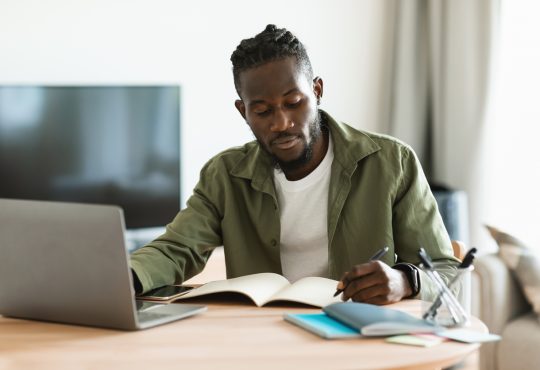 Black man working on laptop and taking notes in notebook
