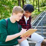Two teens smiling while looking at notebook, leaning on railing against backdrop of trees on campus