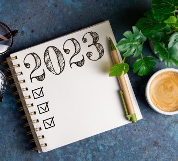 Overhead view of a tabletop with an open notepad with 2023 written and checkboxes underneath it. The table also has a plant, cup of coffee and glasses on it.