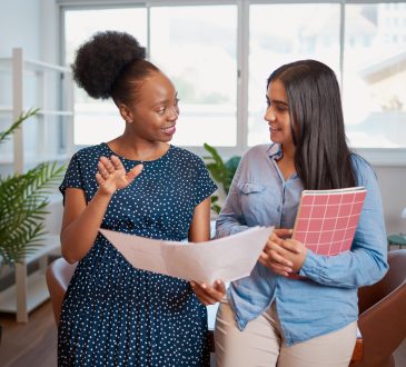 Two women standing next to each other and facing one another. they are talking to each other with one woman holding a piece of paper in front of them.