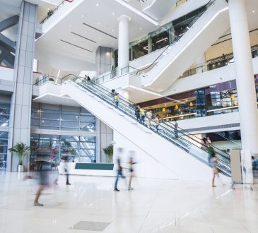 The image shows the interior of a modern, spacious shopping mall with multiple levels connected by escalators, where people are moving about, creating a sense of activity and movement.