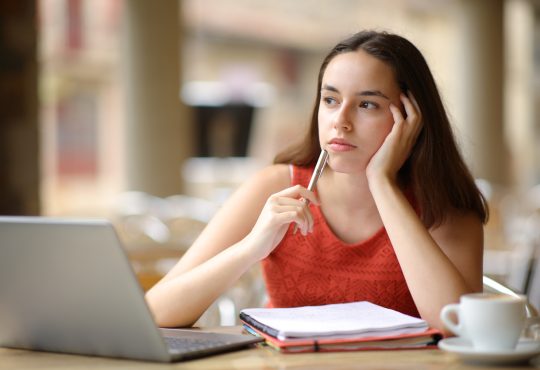 Student sitting at laptop thinking with hand holding side of head and pen raised to mouth