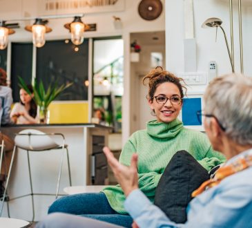 A young woman with glasses and a green turtleneck, smiling and engaging in conversation with an older woman in a modern, brightly lit living space.