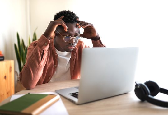 The image shows a young man sitting at a desk, focused on his laptop with a frustrated or concentrated expression, hands touching his head.
