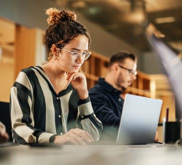 A focused young woman working on a laptop in a shared office space, with other people in the background also engaged in their work, creating a collaborative work environment.