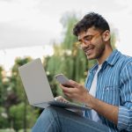 Young man sitting outside with computer balanced on lap, looking at phone in hand