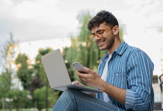 Young man sitting outside with computer balanced on lap, looking at phone in hand