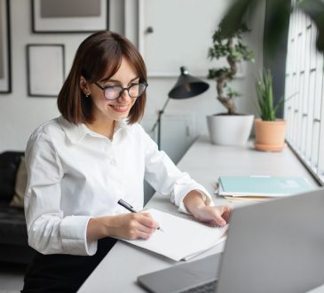 Smiling businesswoman using laptop and taking notes at workplace