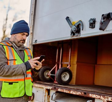 Delivery truck driver standing behind box, checking phone
