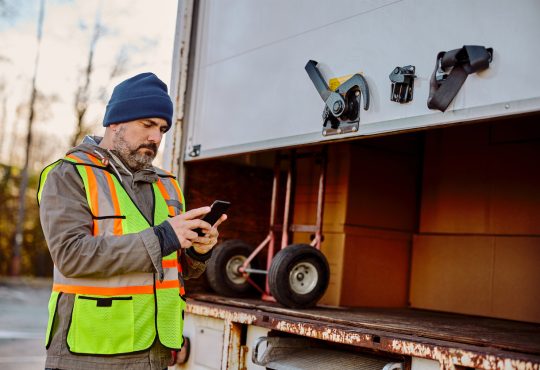 Delivery truck driver standing behind box, checking phone