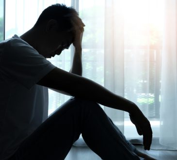 Man silhouetted sitting beside window with head in hands