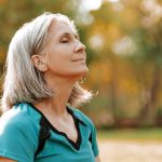 Senior woman wearing sportswear is enjoying a sunny autumn day in the park, closing her eyes and taking a deep breath of fresh air.