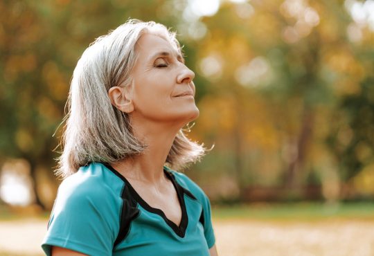Senior woman wearing sportswear is enjoying a sunny autumn day in the park, closing her eyes and taking a deep breath of fresh air.