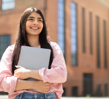 Smiling young woman holding laptop outside of school