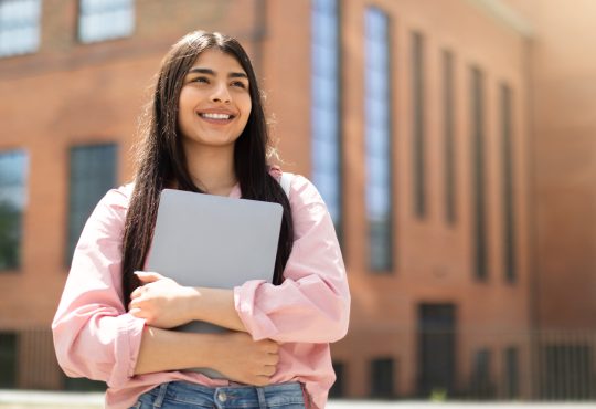 Smiling young woman holding laptop outside of school