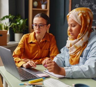 Caucasian young adult woman and Middle Eastern young adult woman wearing hijab learning together in an office, using laptop and taking notes with pens and notebooks