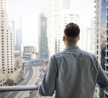 Rear view of young man standing on balcony, gazing out at modern urban landscape filled with high-rise buildings and busy roads, taking moment to admire big city view from his new, luxury apartment