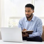 Serious African student man watching online lecture, training seminar on laptop at home, writing notes, summary of webinar, studying on Internet, doing learning homework, speaking on video call