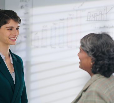European Caucasian young adult gen z diverse business woman lady standing talk workday task indoor job office place. Teen female worker boss manager make conversation give advice helping consulting discuss client insurance benefit