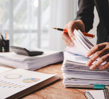 A desk with piles of reports being handled by a set of hands holding a pen. The desk is by a well-lit window, and one pile of reports has a calculator on it.