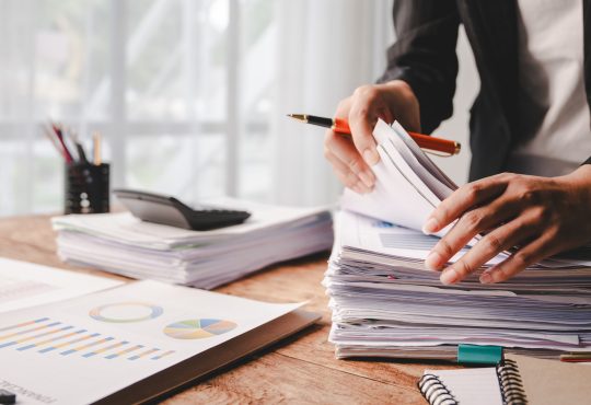 A desk with piles of reports being handled by a set of hands holding a pen. The desk is by a well-lit window, and one pile of reports has a calculator on it.