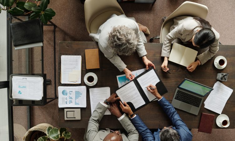 Group of middle aged multiethnic business professionals collaborating around table, reviewing documents and using laptop, top view showing teamwork and corporate meeting environment